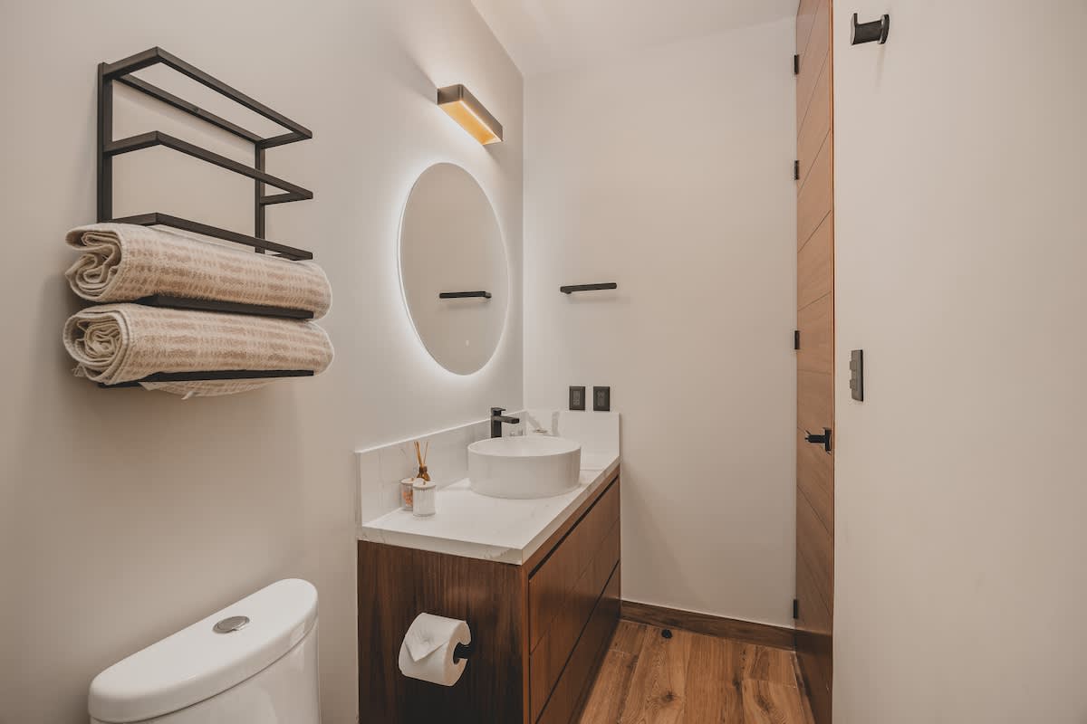A minimalist bathroom features a floating wooden vanity with a white vessel sink and a backlit circular mirror, alongside a toilet and a black metal towel rack with neatly folded towels. The room has light walls and dark wooden flooring.
