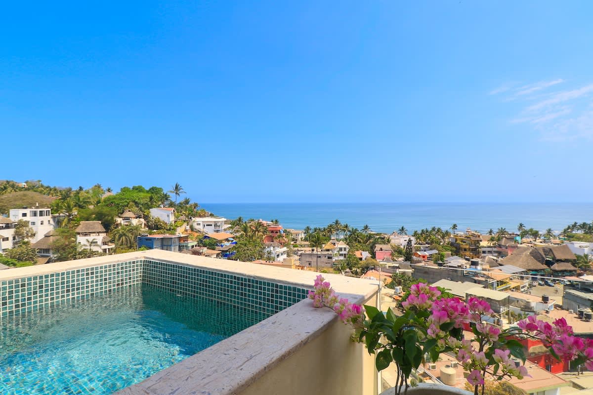 Elevated plunge pool with turquoise mosaic tiles, providing breathtaking panoramic views of the ocean, coastline, and the colorful rooftops of the town below. Pink bougainvillea adds a vibrant touch.