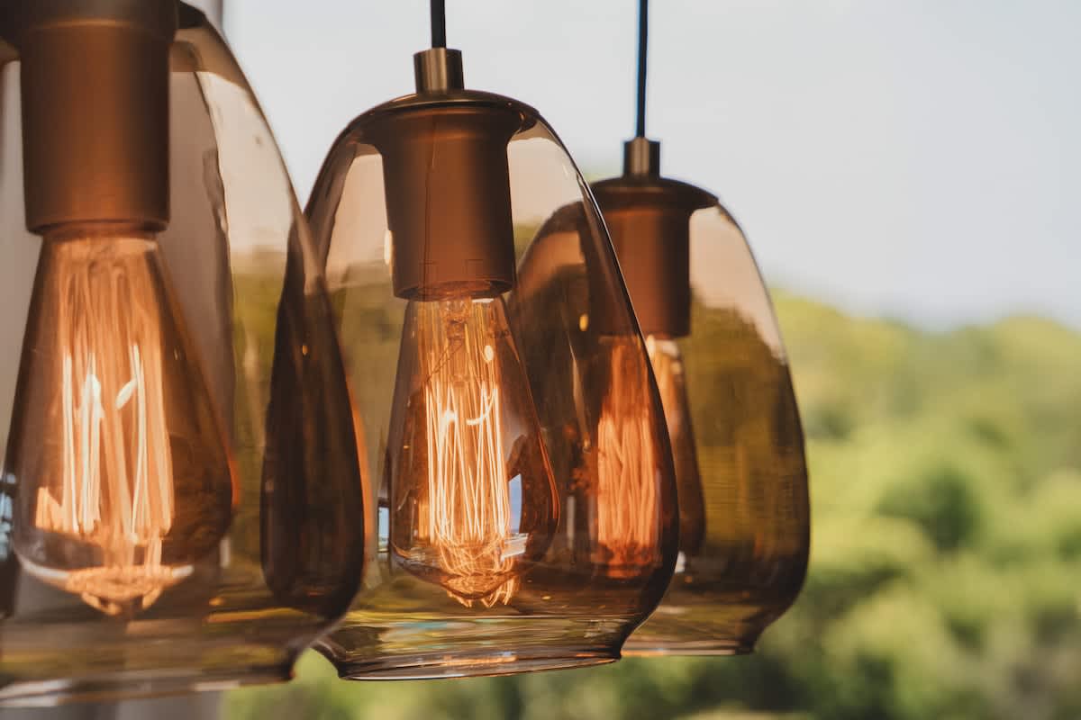 A close-up view of three modern pendant lights with amber-tinted glass shades and exposed filament bulbs, providing warm illumination. A blurred green outdoor landscape is visible in the background.