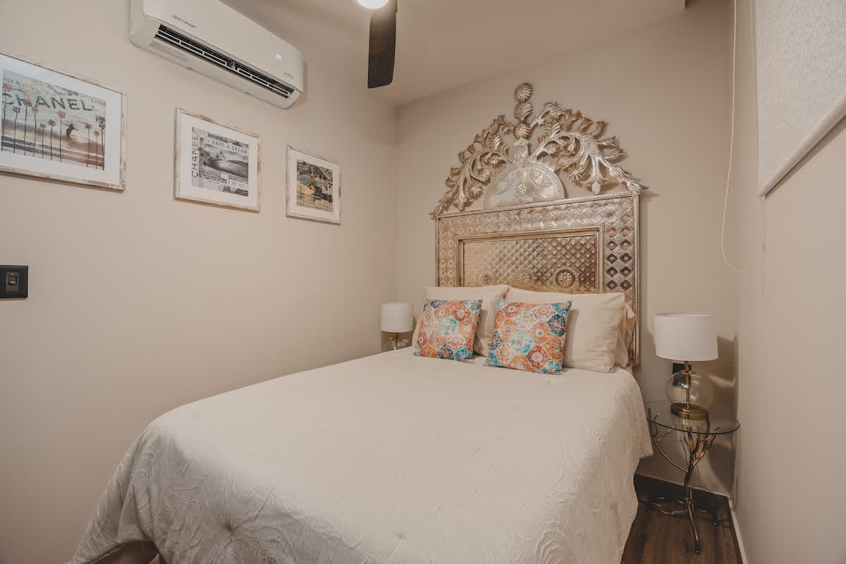 Bright bedroom with a queen-sized bed, an ornate silver headboard, and colorful pillows, featuring an air conditioning unit mounted above the bed and framed artwork on the wall.