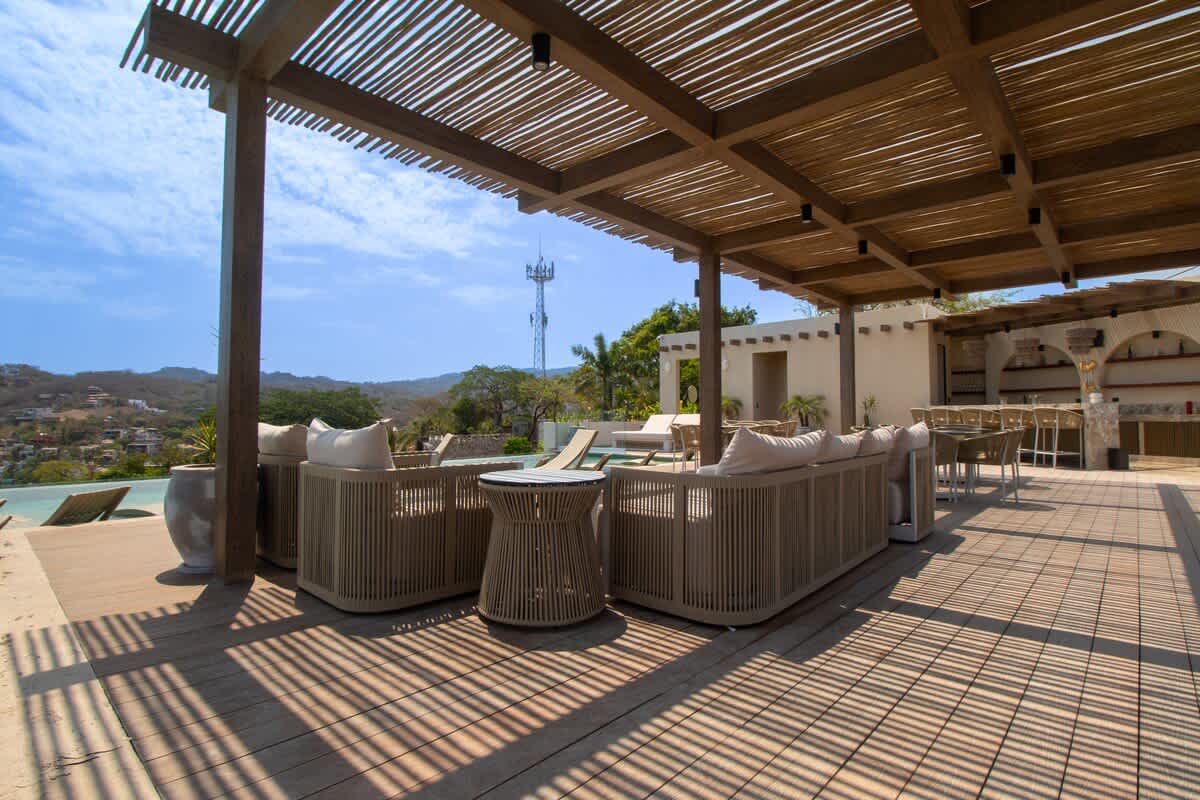 An expansive outdoor patio features comfortable woven lounge seating under a slatted wooden pergola, with a swimming pool visible in the background and lush hills under a bright sky.