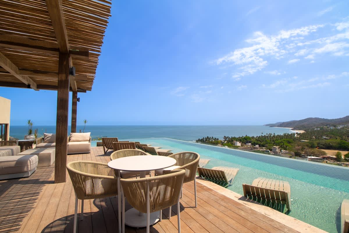 Wide shot of a luxurious outdoor deck area with a large infinity pool offering panoramic ocean and coastline views, featuring a shaded dining table with chairs and additional lounge seating.