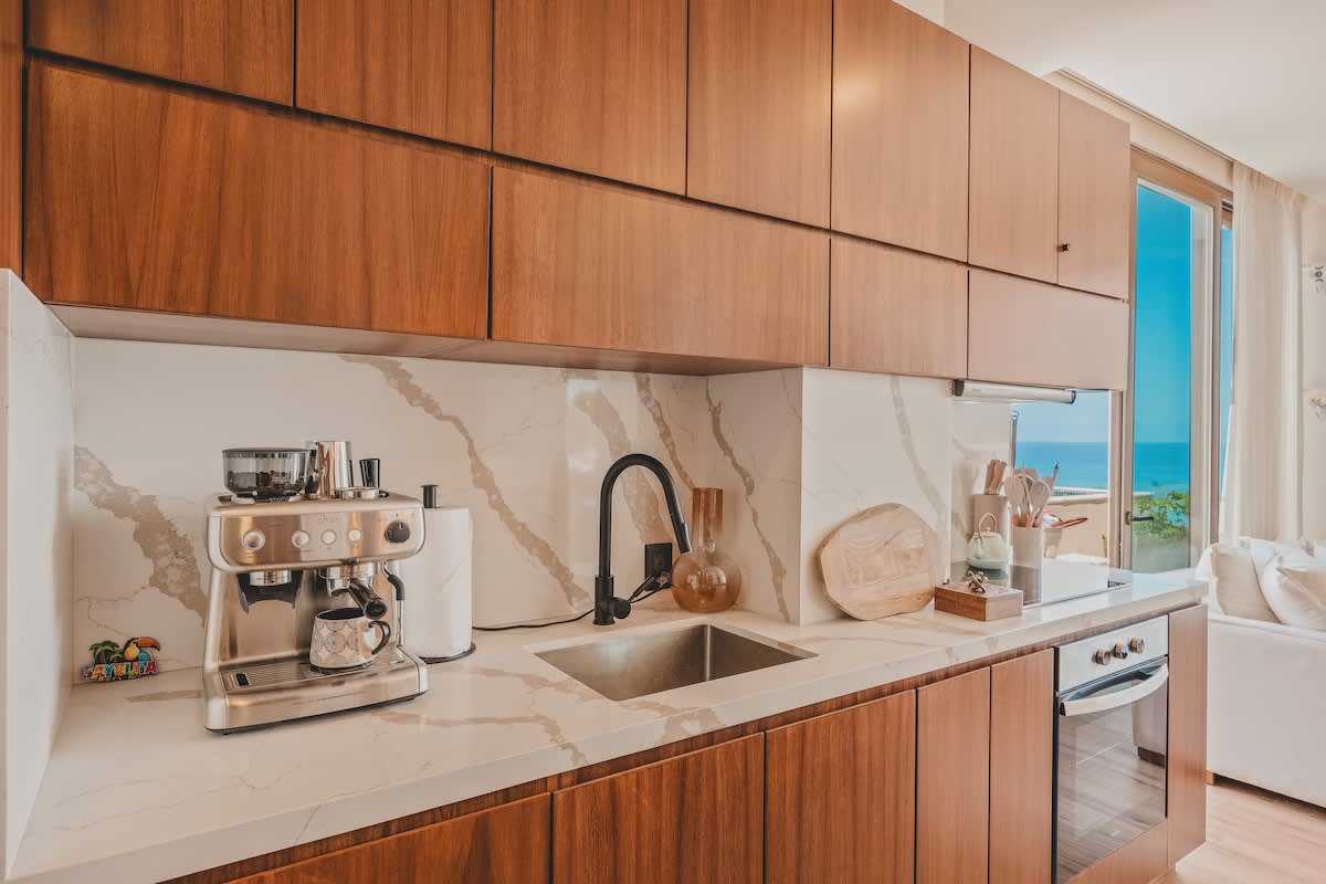 Close-up of a modern kitchen counter with white marble, showcasing a stainless steel espresso machine, a black faucet over a sink, and a view of the ocean through a large window in the background.