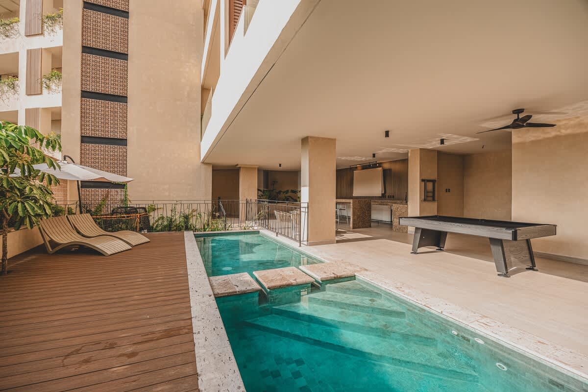 A long, narrow swimming pool with a shallow end is bordered by a wooden deck with lounge chairs and a tiled area featuring a pool table, all under the shade of a modern building. Lush tropical foliage and a patterned building facade are visible in the background.