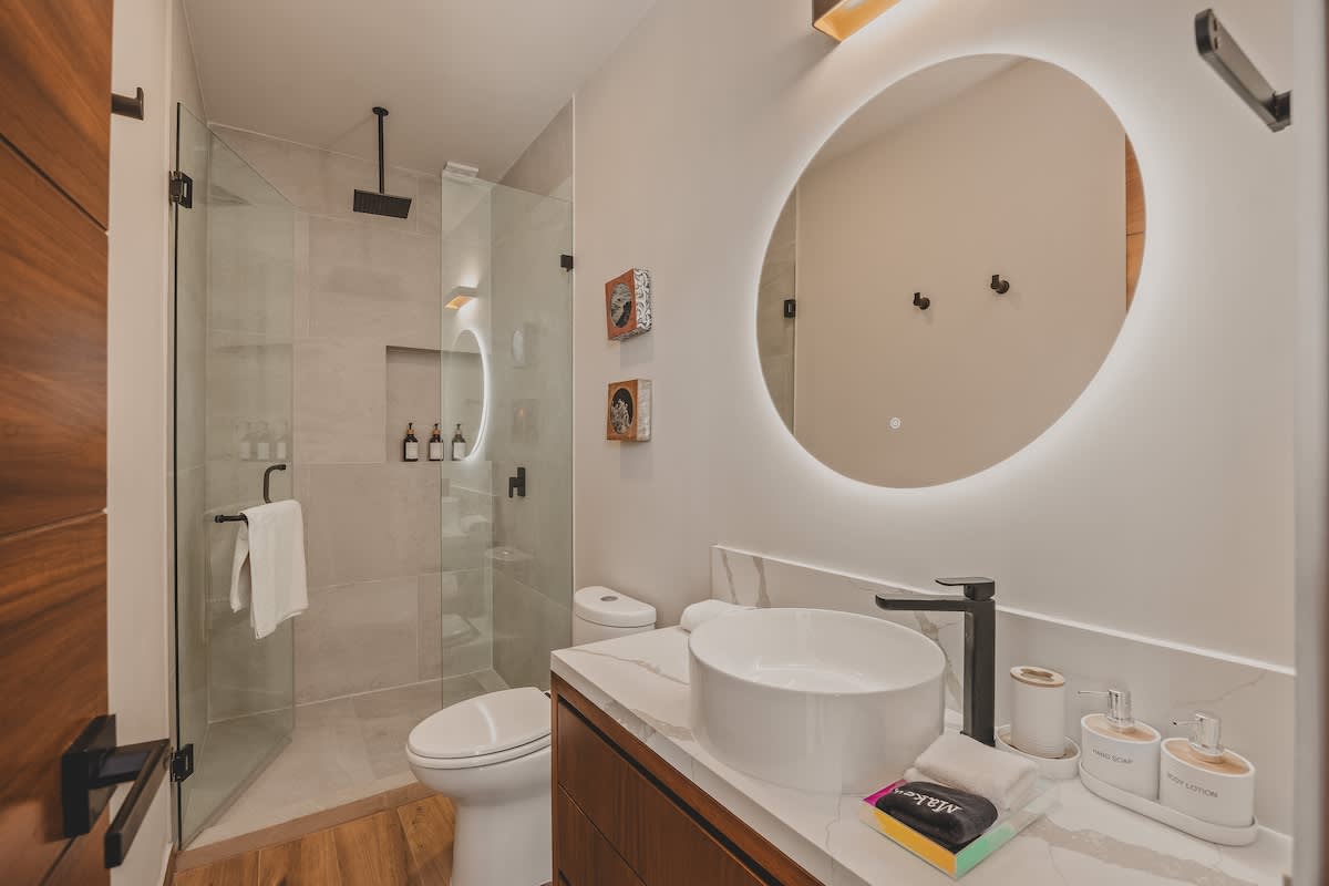 Modern bathroom featuring a glass-enclosed walk-in shower with a rain showerhead, a white vessel sink on a marble-topped vanity, and a large backlit circular mirror. Dark wood accents and minimalist decor complete the space.