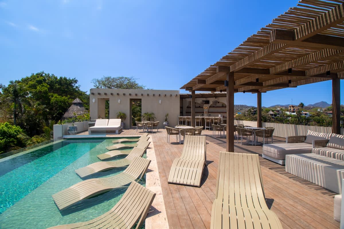 Expansive rooftop deck with an infinity pool featuring in-water lounge chairs, surrounded by a wooden deck with shaded seating areas and a bar, all overlooking a lush landscape and distant town.