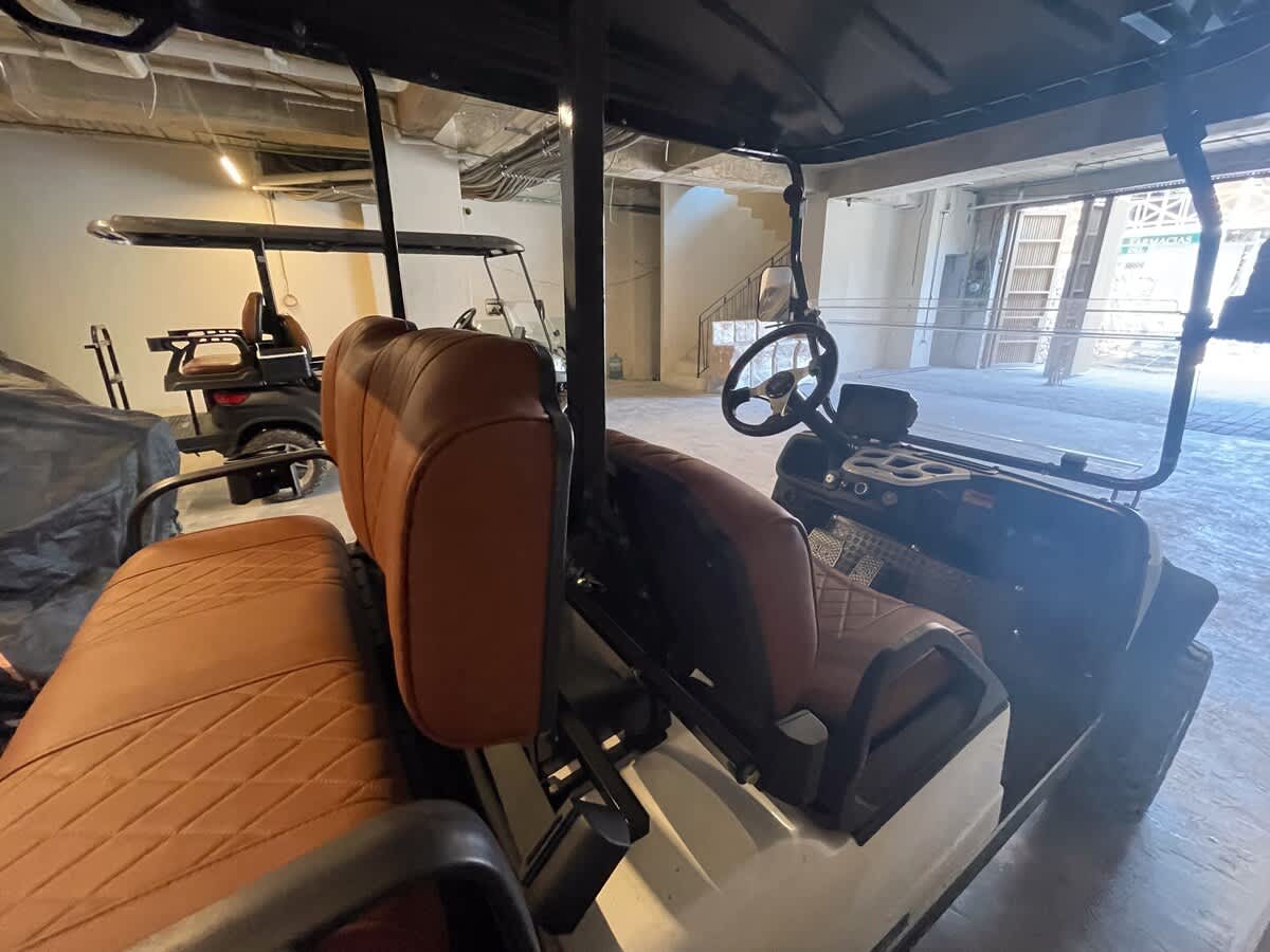 The interior of a golf cart is visible, highlighting the brown quilted seats, steering wheel, and dashboard, with another golf cart parked in the background within a concrete garage.