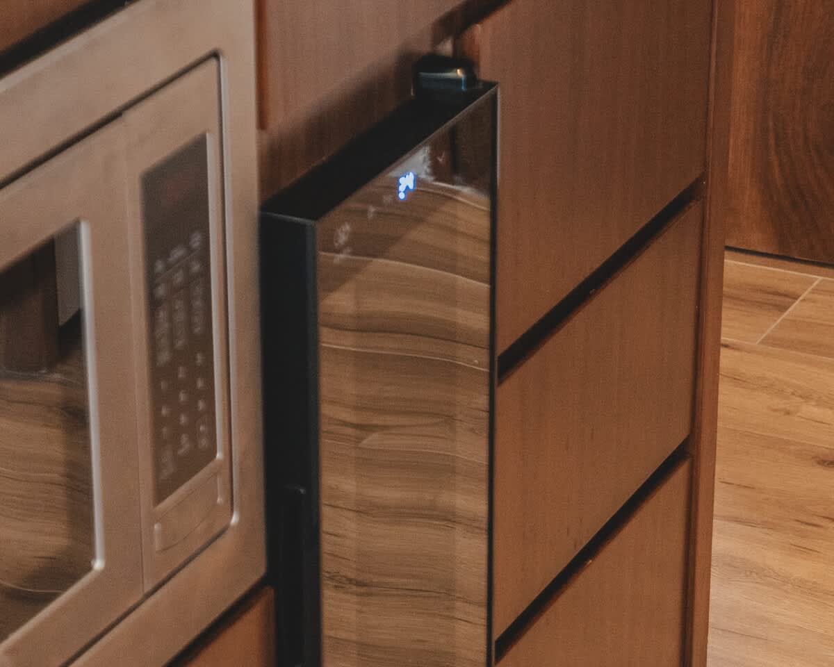 A modern kitchen setup features a stainless steel microwave oven seamlessly integrated next to a sleek, dark-paneled wine cooler or mini-fridge, all built into warm wooden cabinetry.