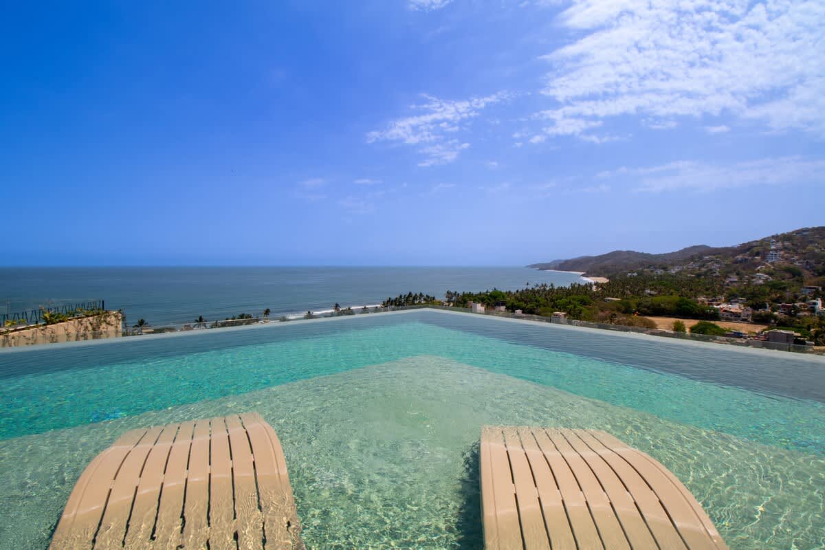 Breathtaking view of an infinity pool seamlessly blending with the horizon, overlooking a vibrant coastal town and the expansive blue ocean under a partly cloudy sky. Two in-pool lounge chairs are visible in the foreground.