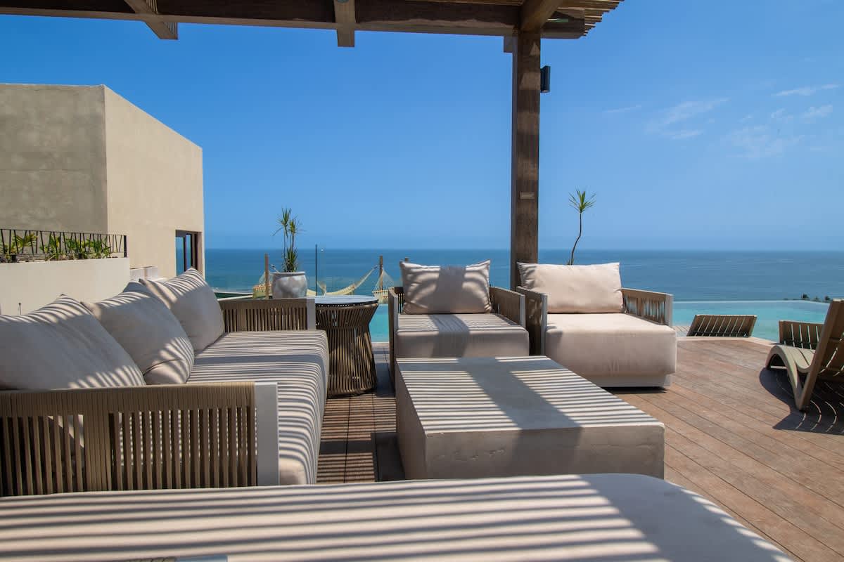 Stylish outdoor lounge area on a wooden deck, featuring comfortable light-colored sofas and armchairs under a slatted pergola, with a stunning backdrop of the ocean and distant hills.