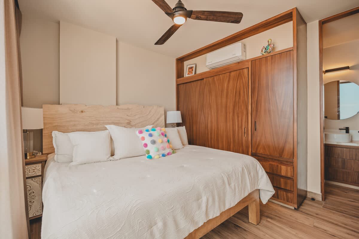 Bright bedroom featuring a large bed with a light wood headboard, white bedding, and a colorful decorative pillow, complemented by a dark wood ceiling fan and built-in wooden cabinetry with an air conditioning unit above.