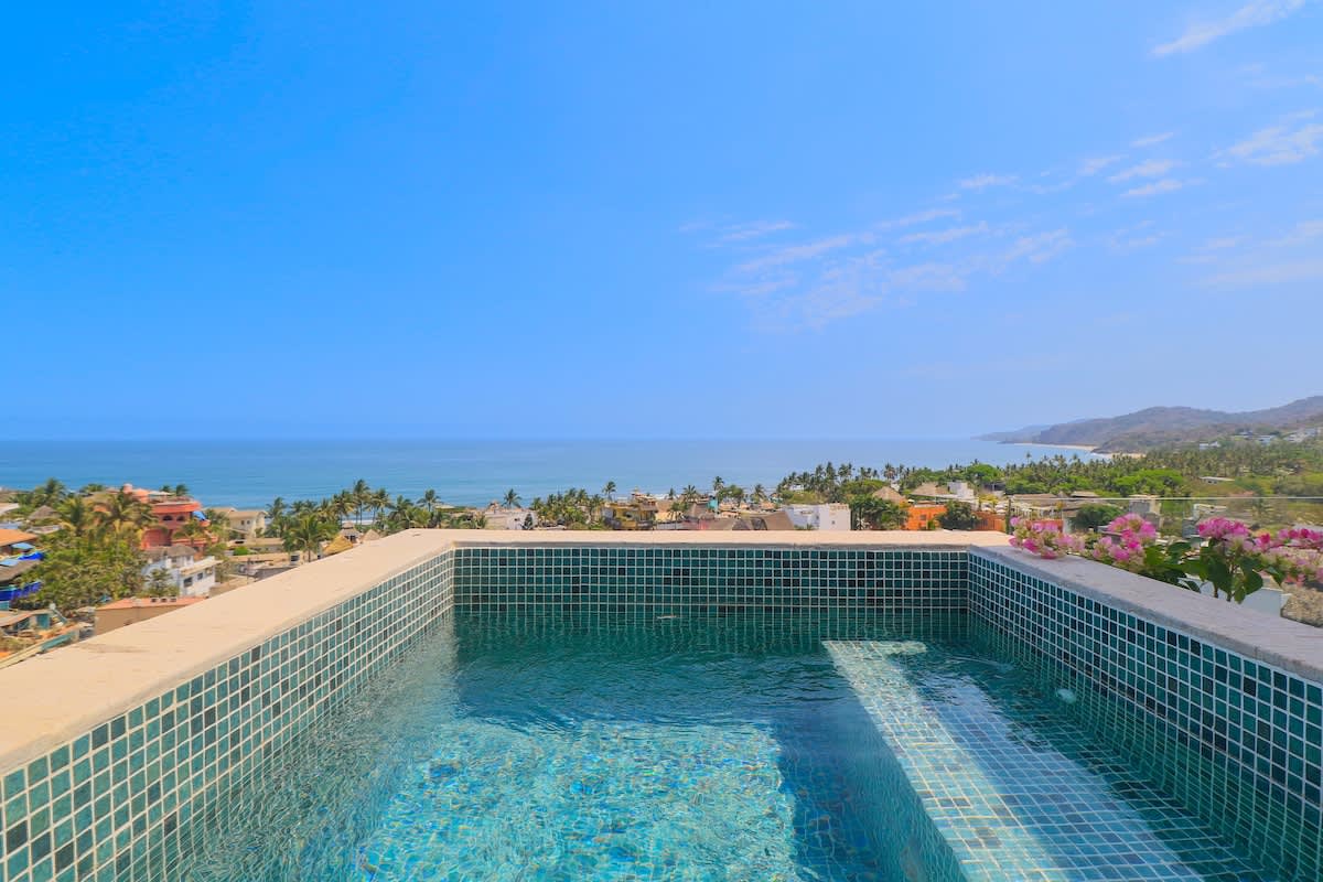 Close-up view of the sparkling blue-tiled plunge pool, set against a backdrop of the vast blue ocean and a distant coastline. The clear water invites relaxation with stunning natural scenery.