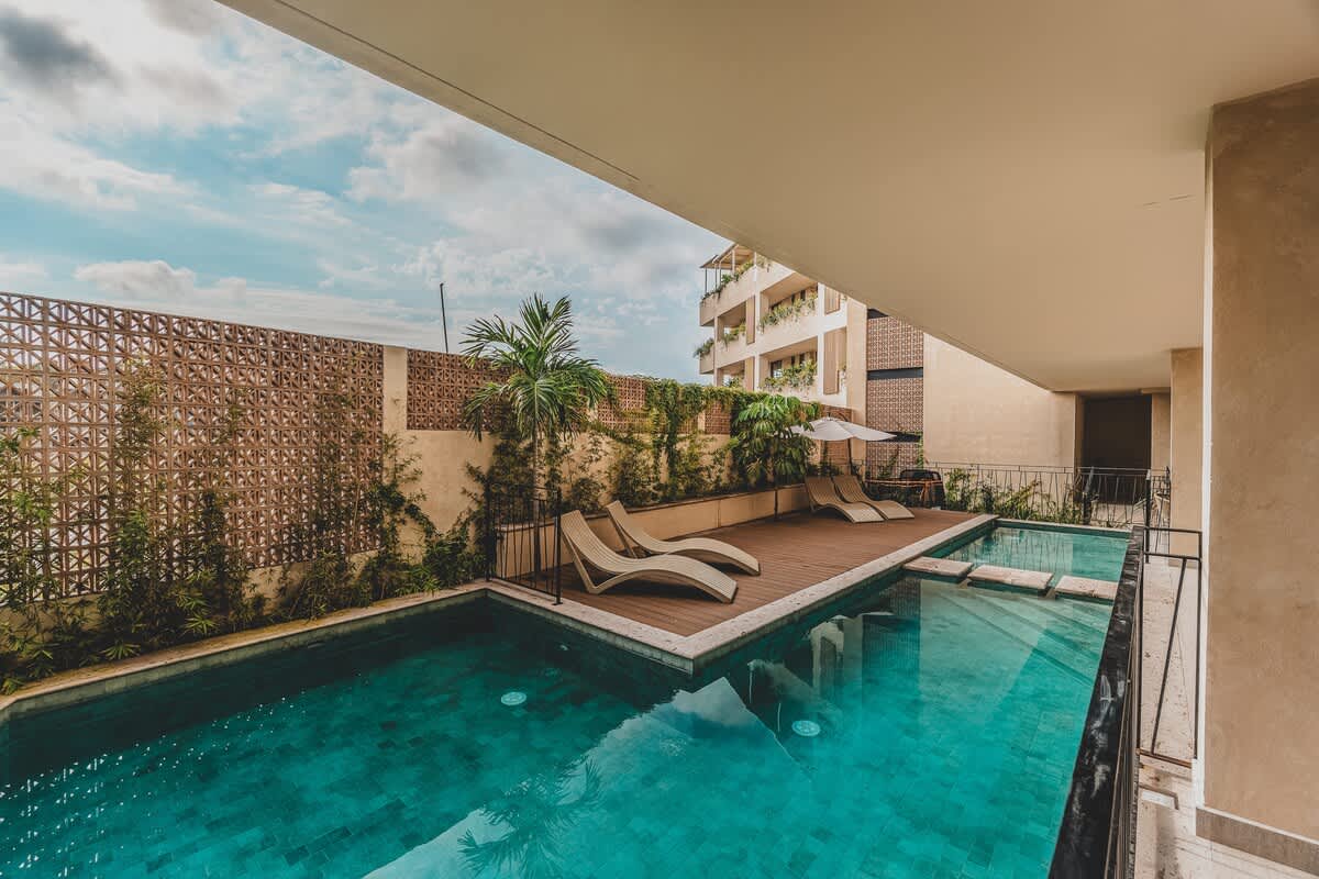 A serene swimming pool with clear turquoise water, bordered by a wooden deck with two contemporary lounge chairs, and a decorative wall covered in climbing vines, under a bright sky.