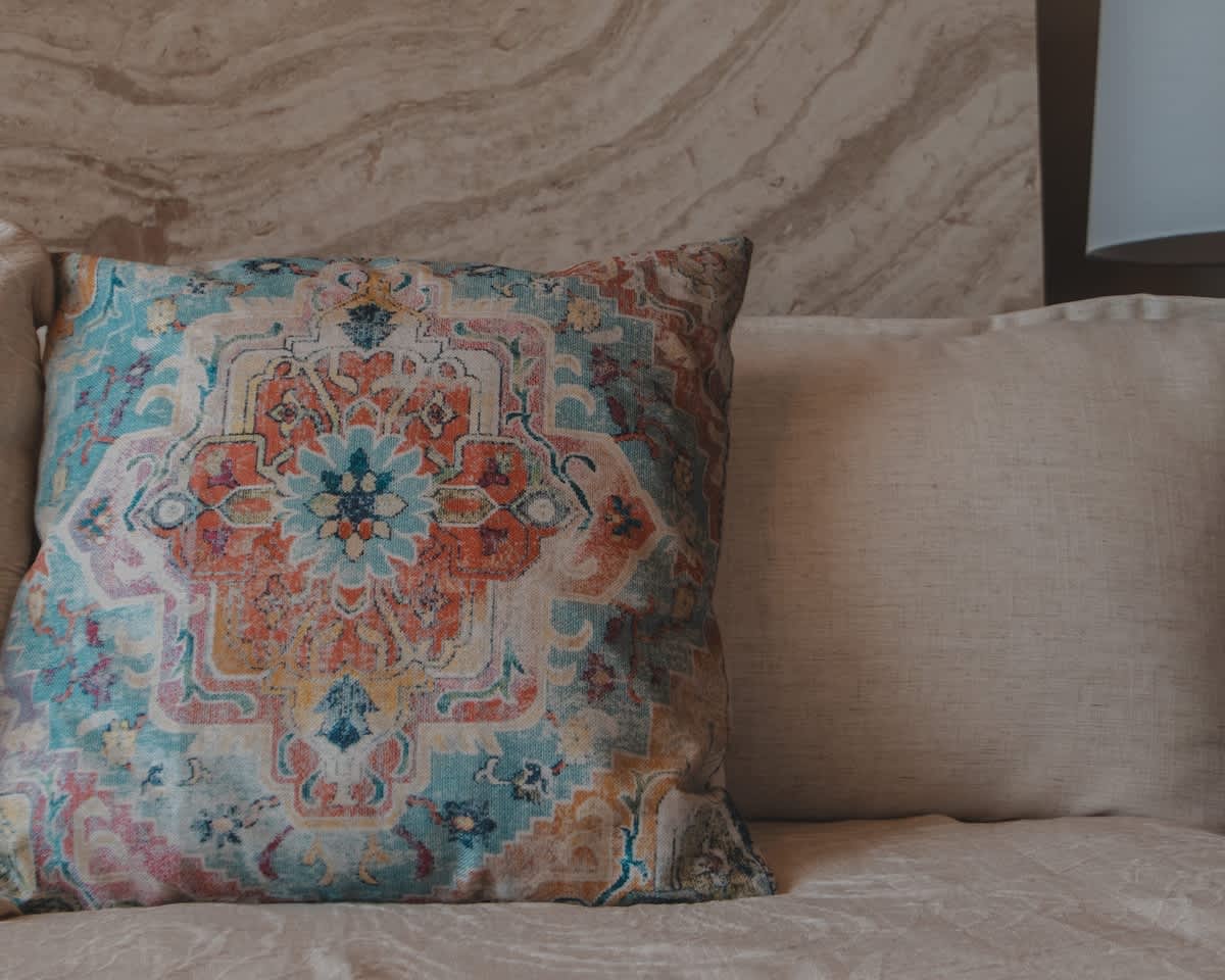 A vibrant decorative pillow with an intricate blue, orange, and white mandala-like pattern rests on a light-colored bed or sofa. A subtle marble-patterned wall is visible in the background.