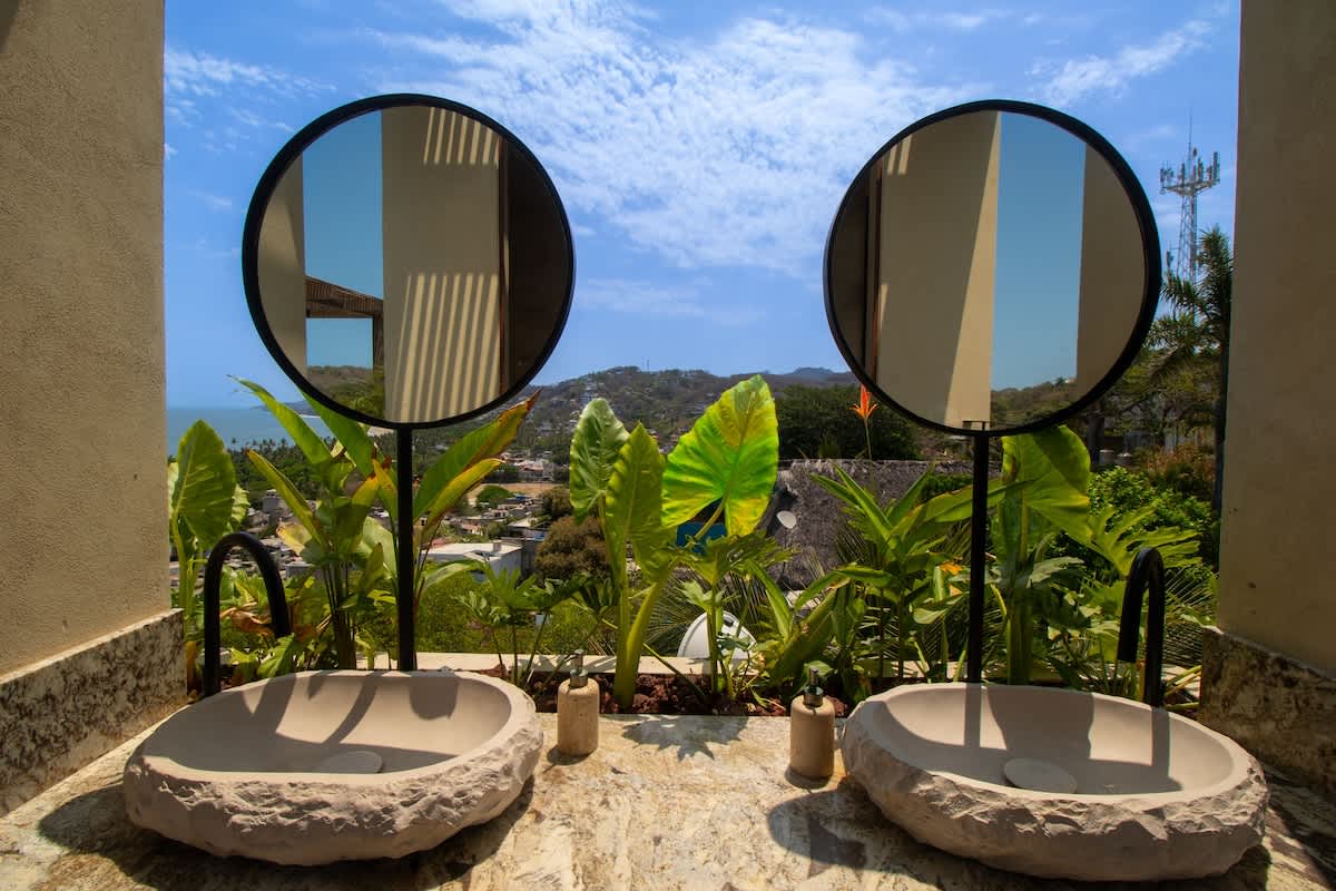 A luxurious outdoor-facing bathroom features two rough-hewn stone vessel sinks and round black-framed mirrors, offering panoramic views of the tropical landscape and ocean beyond. The vanity top is a light-colored, veined stone.
