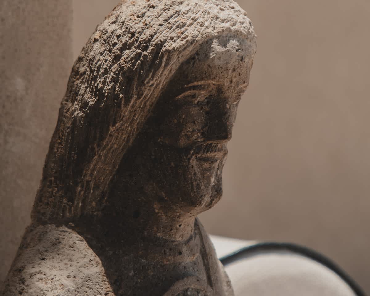 A detailed close-up of a weathered stone sculpture depicts the head and shoulders of a figure, possibly hooded, showcasing rough textures and ancient artistry.