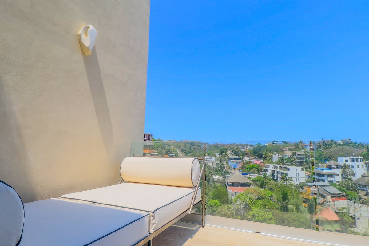 A modern outdoor balcony with a white daybed and glass railing offers a breathtaking, expansive view of a coastal town, lush greenery, and the distant ocean under a clear blue sky.
