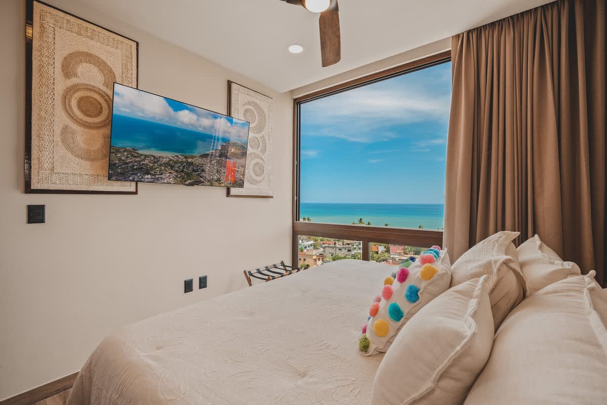 Cozy bedroom with a large bed, a wall-mounted television, and floor-to-ceiling windows offering picturesque views of the ocean and the vibrant town. Decorative wall art complements the serene ambiance.
