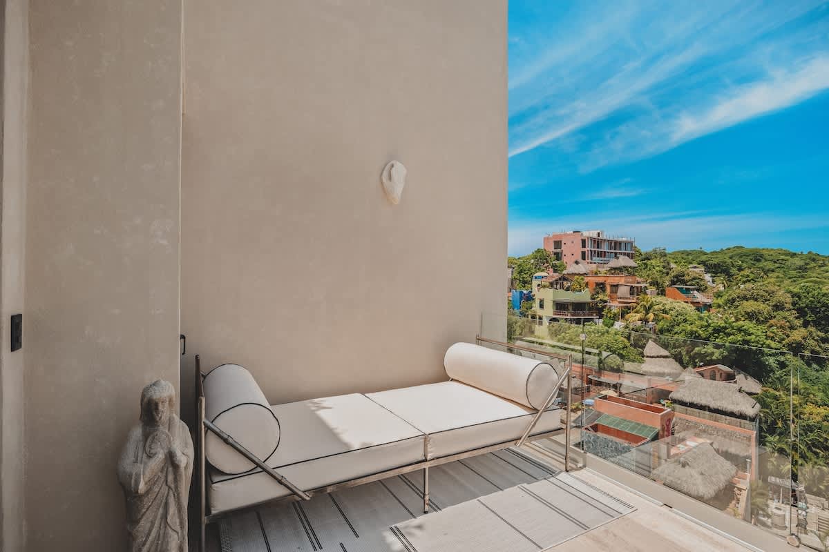 A modern outdoor balcony features a white daybed with black piping, a white statue, and glass railings, offering panoramic views of a vibrant town nestled among green hills under a clear blue sky.