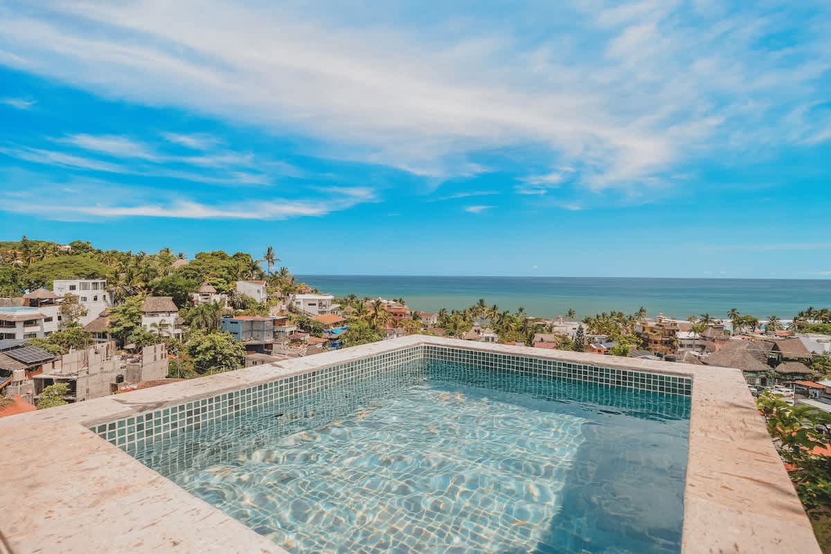 Close-up of a small, tiled rooftop plunge pool with sparkling blue water, offering a stunning panoramic view of the ocean and a vibrant coastal town under a bright blue sky.