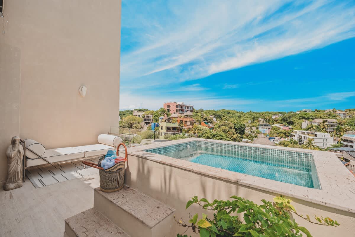 A spacious rooftop deck featuring a small plunge pool, a modern white lounge chair, and lush greenery, all set against a backdrop of a vibrant coastal town and distant hills.
