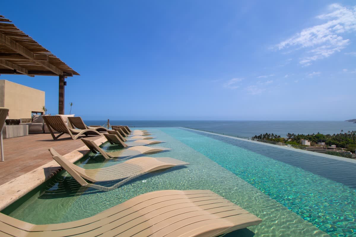 Long infinity pool with multiple in-water lounge chairs, set on a wooden deck under a partial pergola, offering sweeping views of the ocean and a distant coastal landscape.