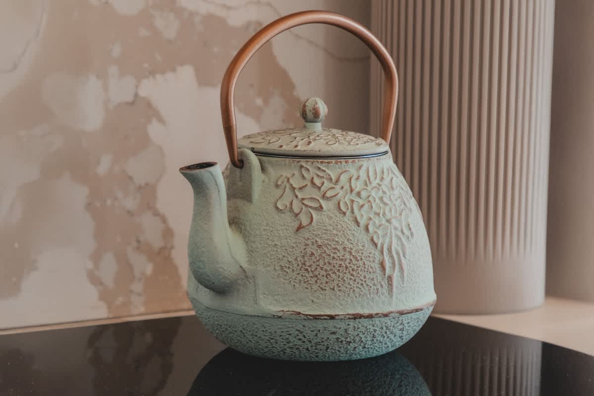 A rustic light blue cast iron teapot with an embossed leaf pattern and a wooden handle rests on a black induction cooktop. A fluted beige vase is visible in the soft-focus background.