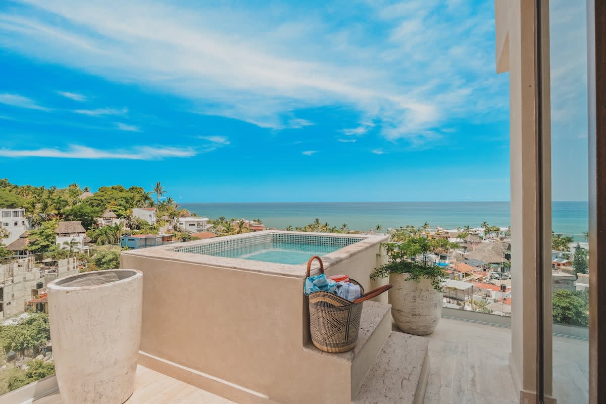 Panoramic view from a rooftop featuring a small tiled plunge pool overlooking a coastal town and the vast blue ocean under a clear sky. A woven basket with towels sits beside the pool.