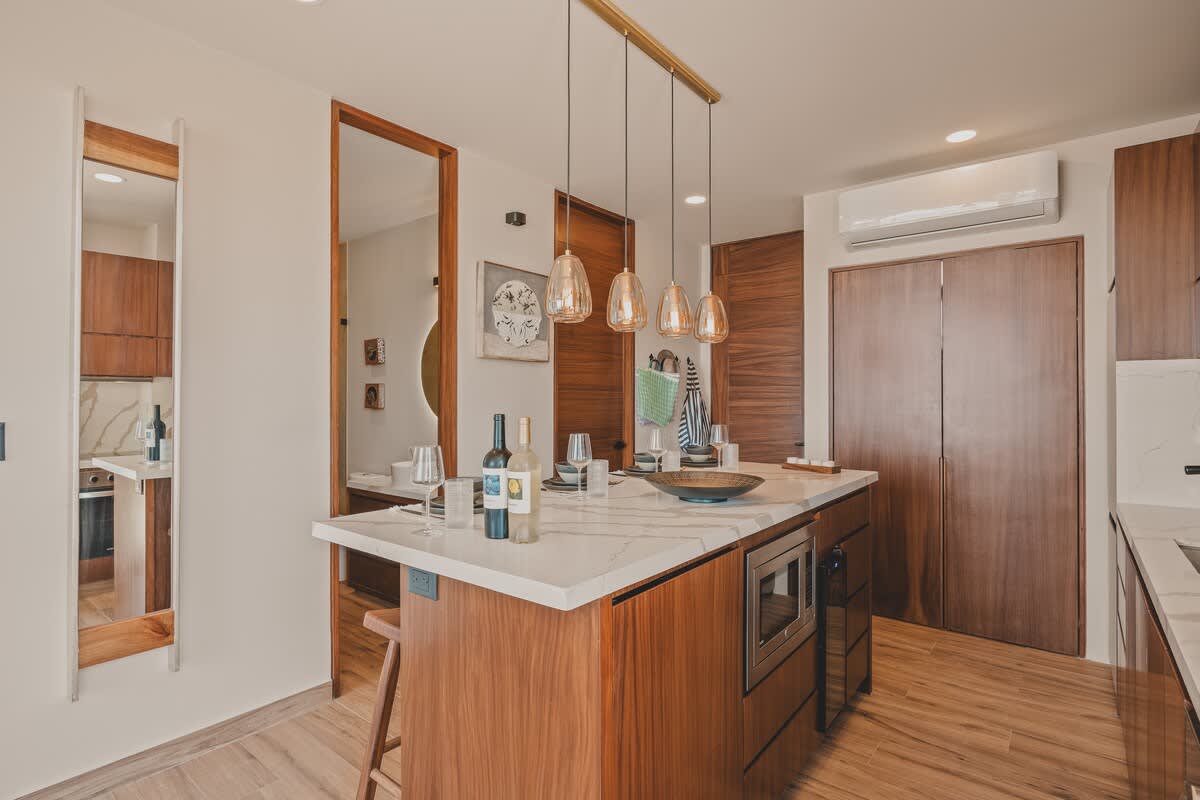 A spacious, modern kitchen showcases a large wooden island with a white marble countertop, pendant lighting, and integrated appliances, leading to a dining area with a mirror. Warm wooden doors and flooring complete the contemporary design.