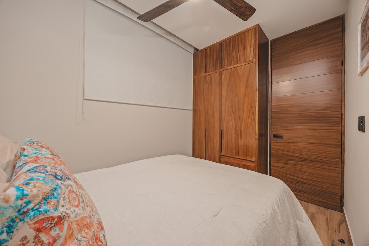 A cozy bedroom features a white-quilted bed with a colorful patterned pillow, set against a light gray wall and a large wooden wardrobe and door. A wooden ceiling fan is visible above.