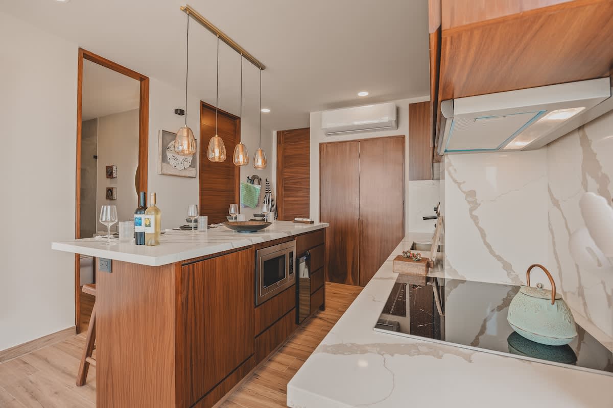 A modern kitchen features a large island with a white marble countertop, integrated microwave, and wine fridge, complemented by warm wooden cabinetry and pendant lights. An induction cooktop and a decorative teapot are visible on the right.