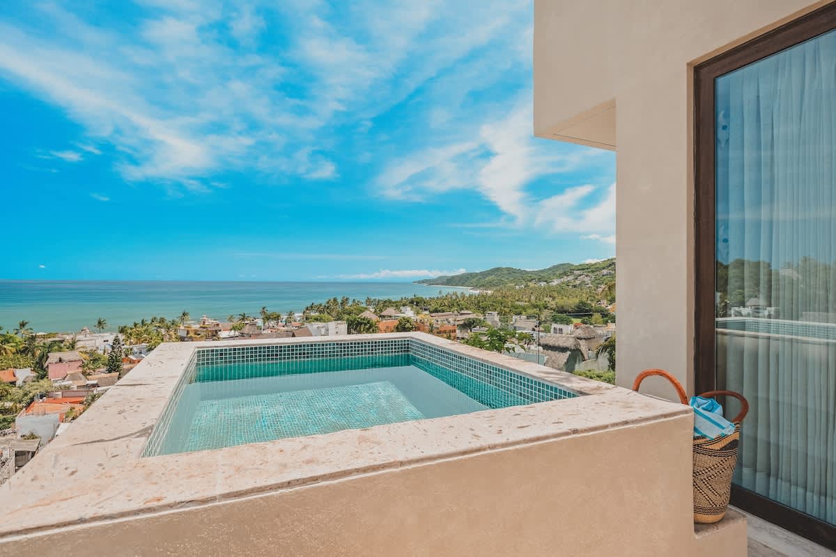 A small, tiled rooftop plunge pool with clear water, offering an expansive view of the ocean coastline, distant mountains, and a bustling coastal town under a bright blue sky.