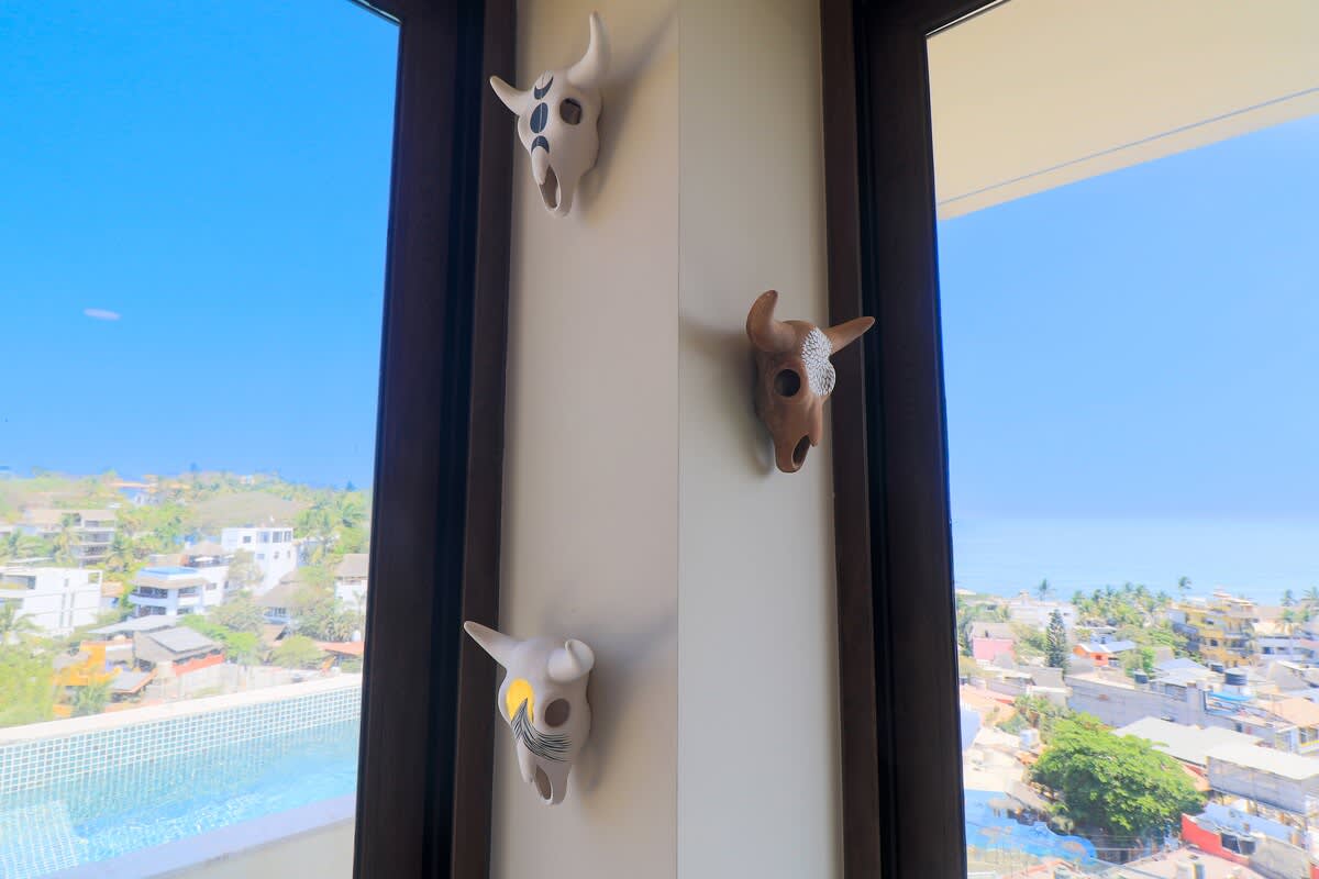 A bright corner of a room features large windows overlooking a vibrant town, the ocean, and a private pool, adorned with three unique, decorative skull-shaped wall hangings.