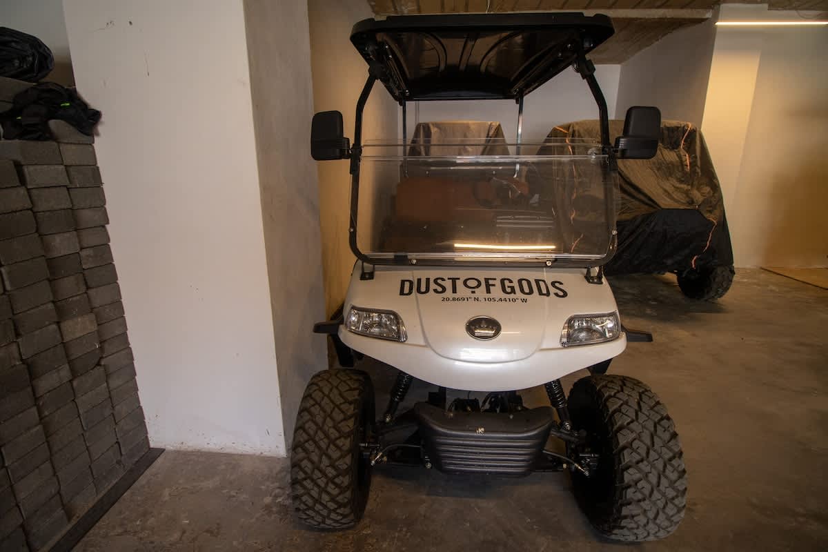 A white golf cart with "DUST OF GODS" branding parked in a concrete garage or storage area, with some building materials stacked on the left.