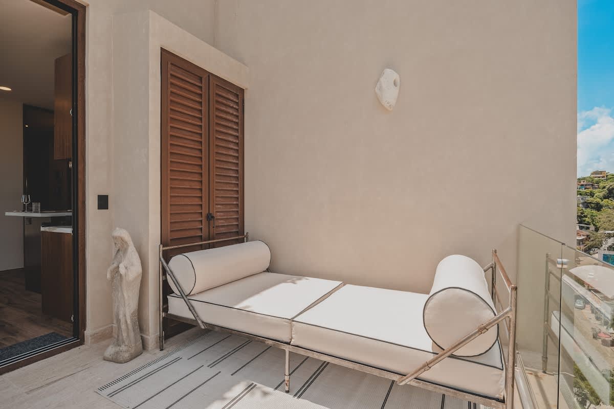 A cozy outdoor balcony with a white daybed and a decorative statue provides a serene lounging space, adjacent to a wooden-shuttered doorway leading indoors, all set against a light-colored wall.