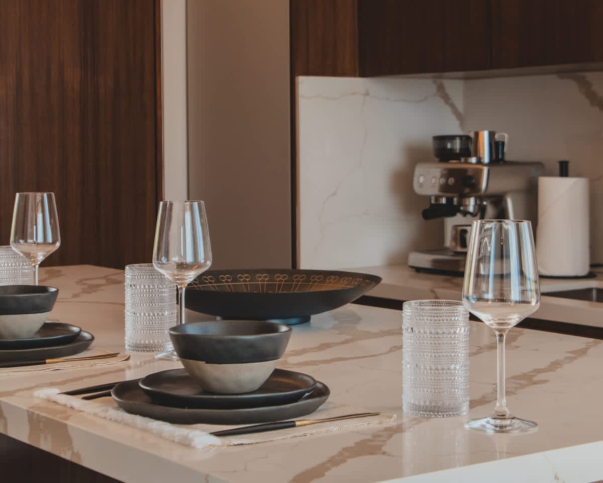 A modern dining setup on a white marble countertop, featuring dark ceramic plates, bowls, and clear wine glasses, with a coffee machine visible in the background against dark wood cabinetry.