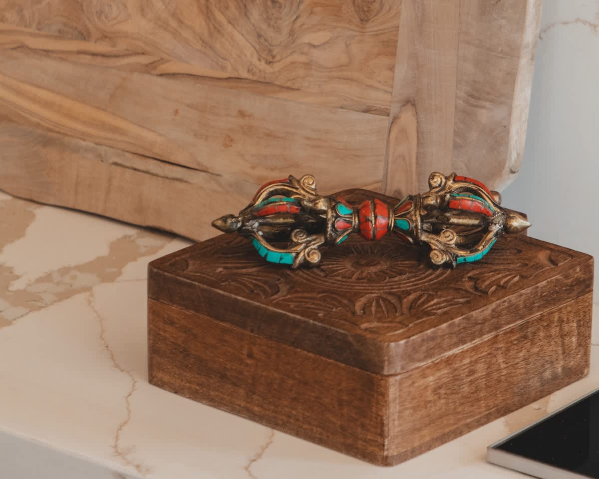 An ornate metal decorative object, possibly a vajra, with intricate red and turquoise gemstone accents, rests elegantly on a richly carved wooden box.