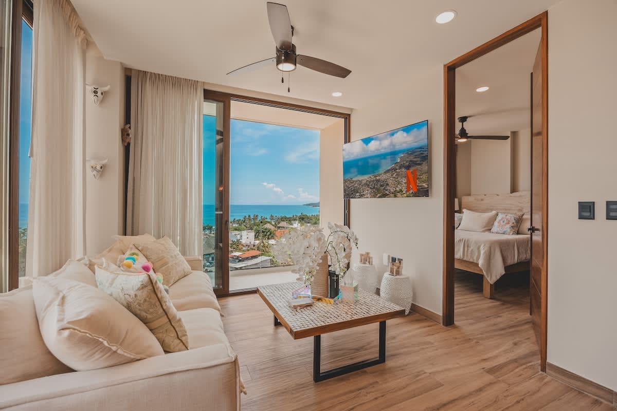 Inviting living room with a cream sofa, mosaic coffee table, and a wall-mounted TV, offering stunning ocean views through large glass doors. An open doorway leads to a bright bedroom.