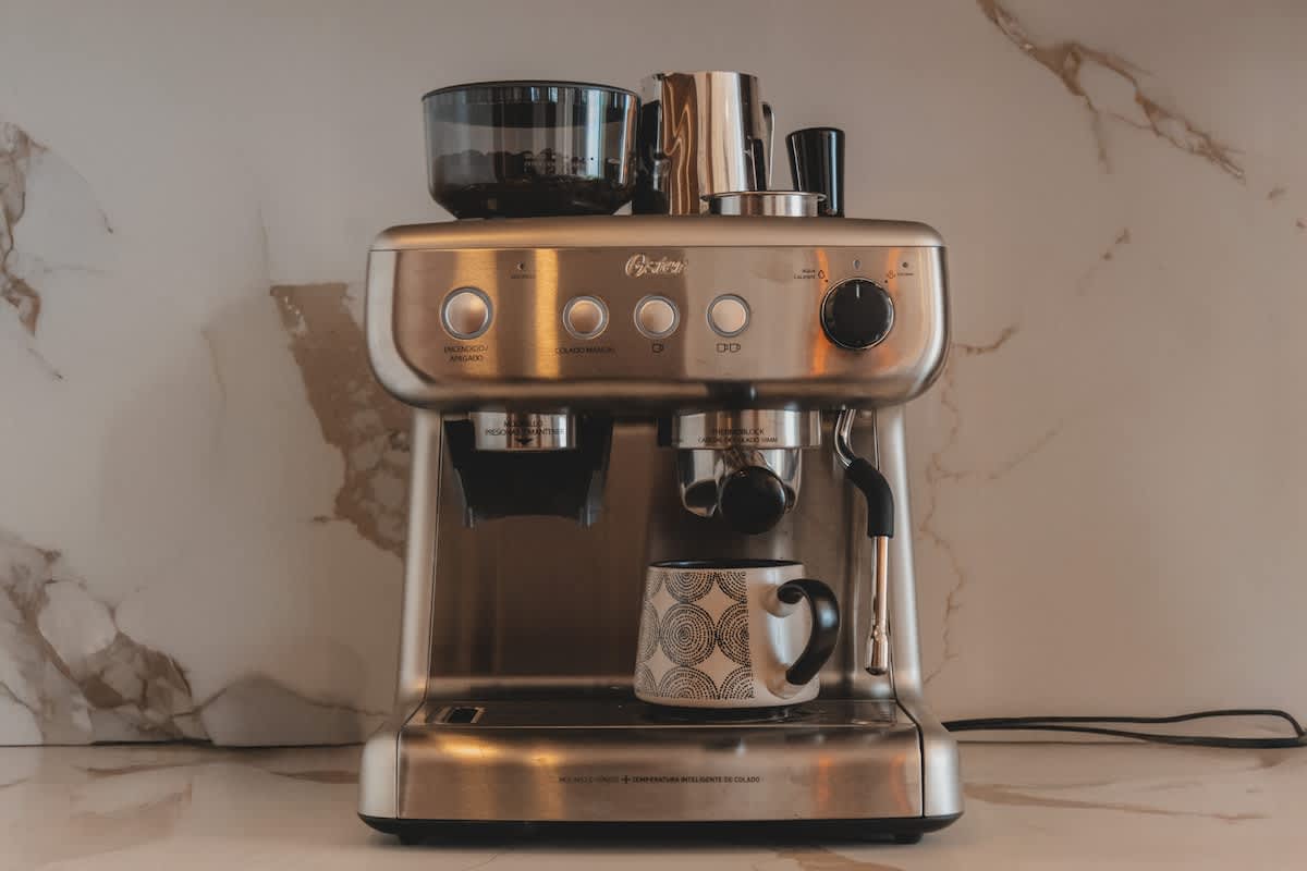A sleek, brushed stainless steel espresso machine with a built-in grinder sits on a white marble countertop, accompanied by a patterned ceramic mug. The background features a subtle marble-patterned wall.
