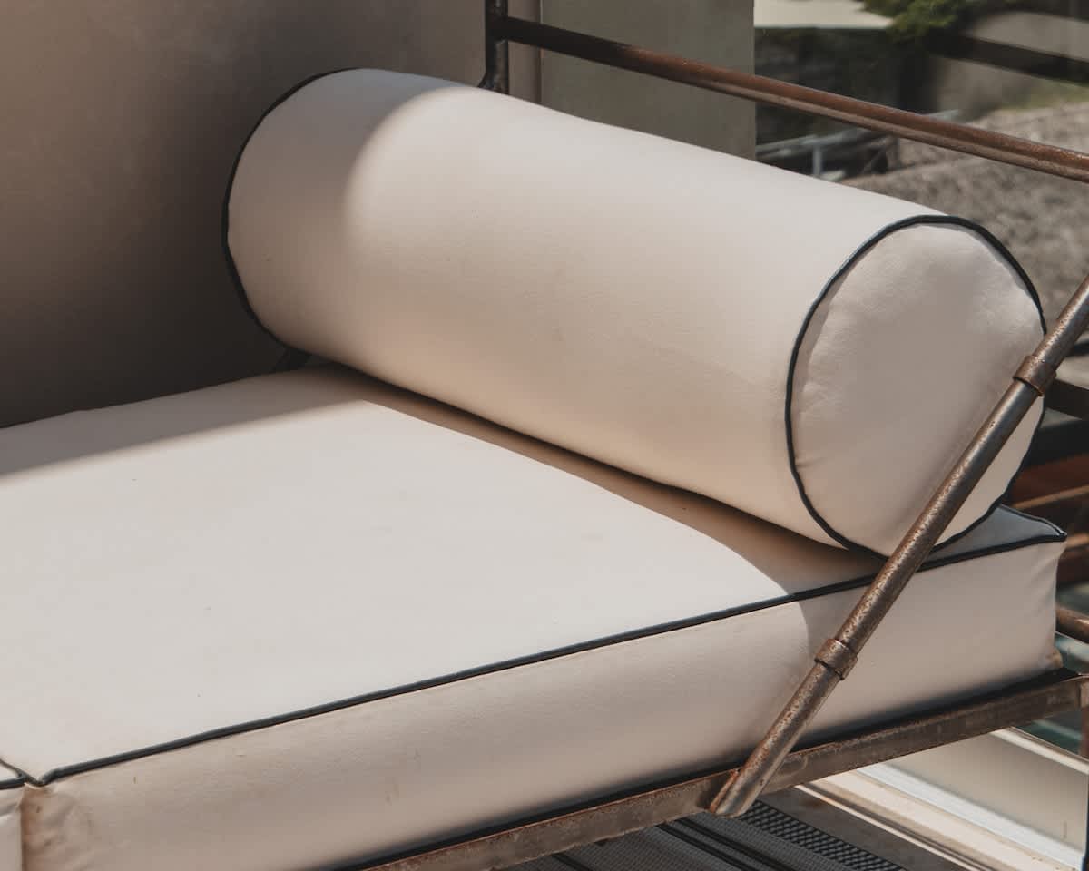 A close-up view of an off-white outdoor cushion with black piping, resting on a rustic metal frame, suggesting comfortable seating on a balcony or patio.