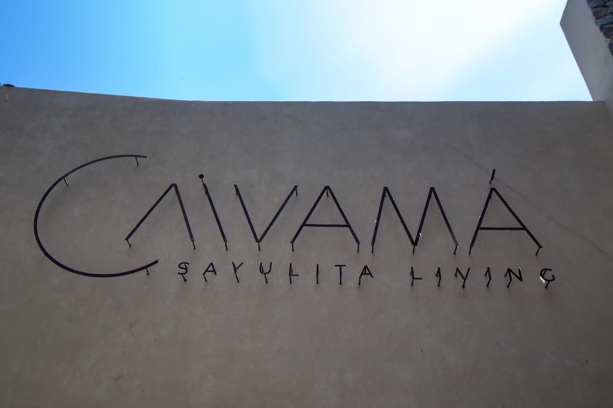 A large, modern sign with metal letters spelling 'CAVAMA SAYULITA LIVING' is mounted on a light-colored stucco wall under a bright blue sky, indicating the property's name.