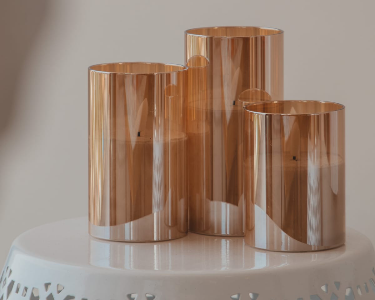 Three elegant amber-colored glass candle holders, varying in height, are artfully arranged on a white, decorative stand with a cut-out pattern.
