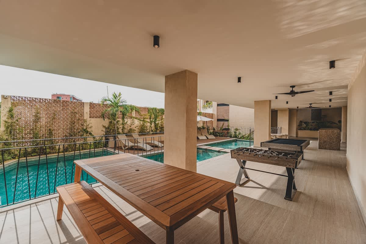 A covered outdoor recreation area featuring a long wooden picnic table with benches, a foosball table, and a pool table, all overlooking a turquoise swimming pool with a black metal railing.