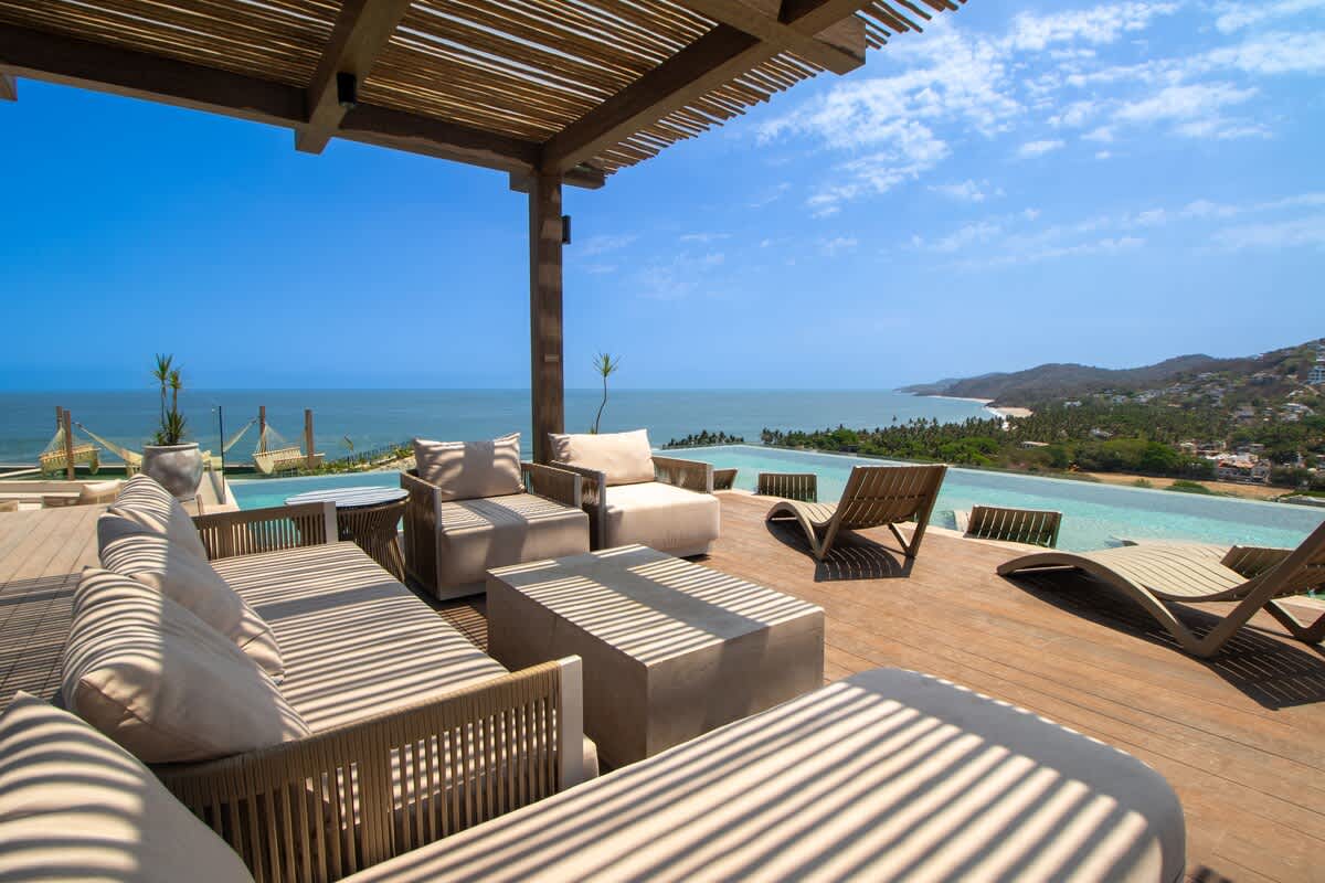 Expansive outdoor deck with a wooden pergola providing shade, furnished with comfortable lounge seating and chaises, overlooking a stunning infinity pool that blends into the vast ocean horizon and a coastal landscape.