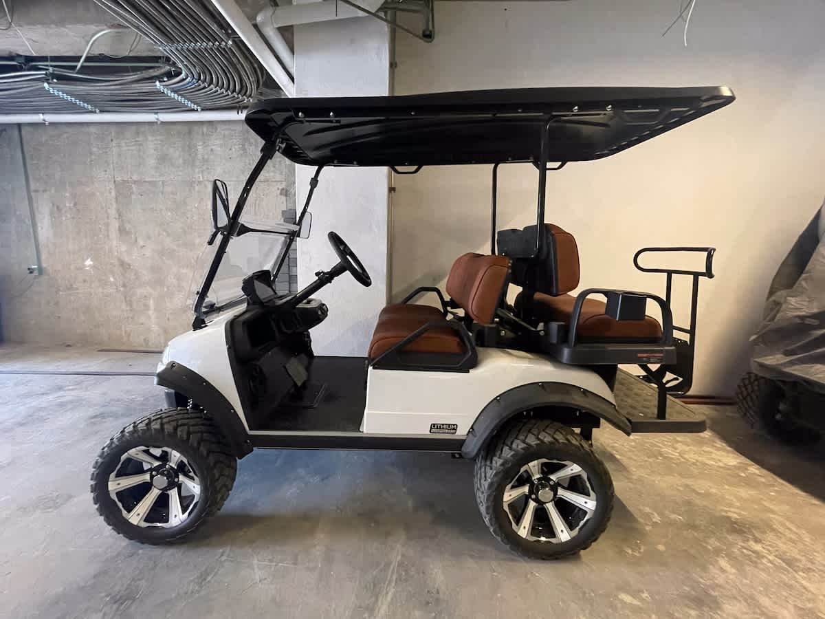 A white golf cart with a black roof and brown quilted seats is parked on a concrete floor, showcasing its large off-road tires and side profile.
