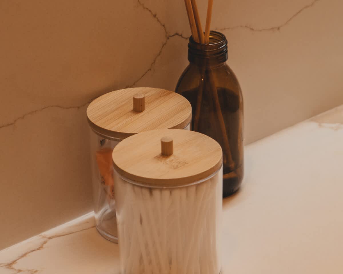Close-up of bathroom amenities on a light marble counter, including two clear jars with bamboo lids holding cotton swabs and pads, and an amber glass diffuser bottle with reeds.