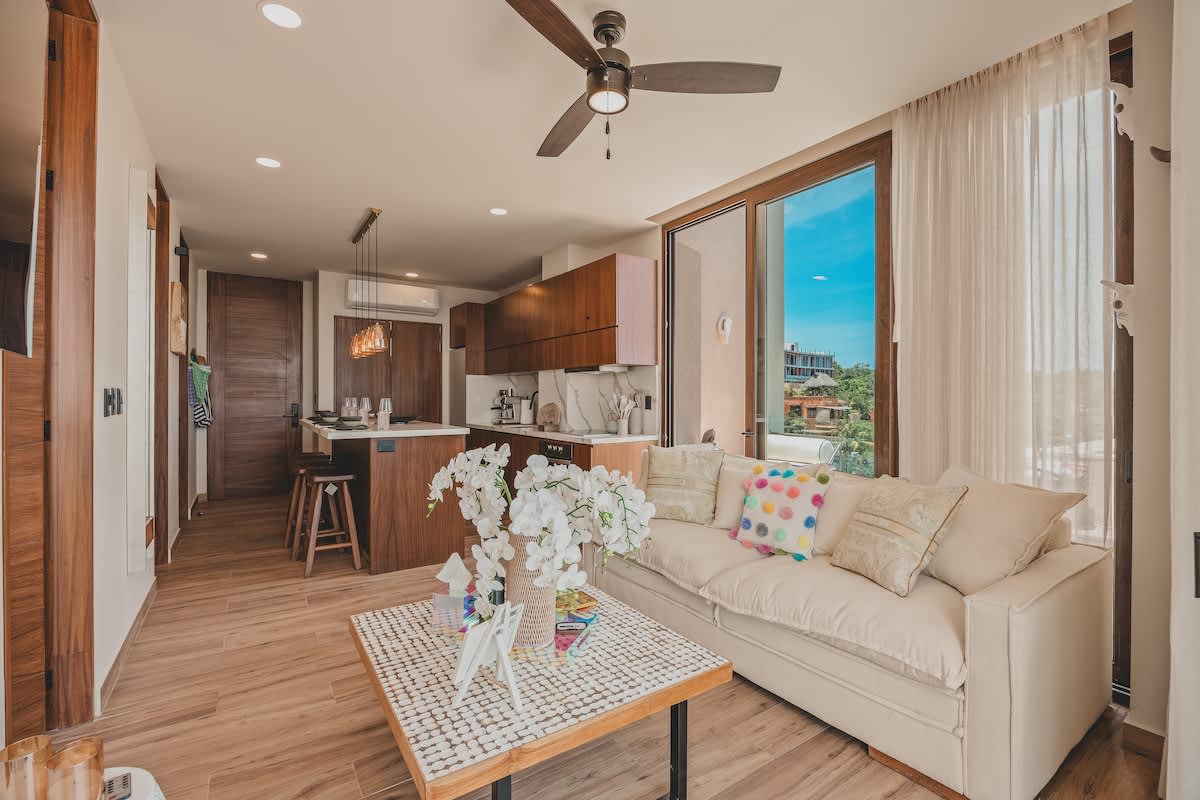 Spacious open-concept living area and kitchen, featuring a cream sofa with colorful pillows and a mosaic coffee table. The modern kitchen with wooden cabinets is visible in the background, all bathed in natural light.