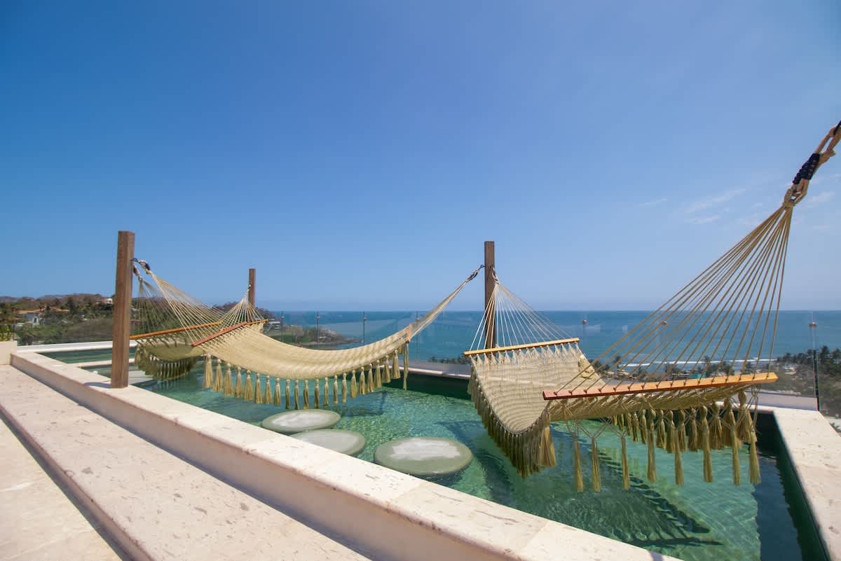 Serene infinity pool with two woven hammocks suspended over the water, offering a unique relaxation spot with panoramic views of the vast blue ocean and distant coastline under a clear sky.