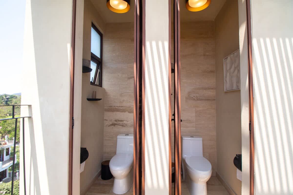 Two individual toilet stalls with modern white toilets are visible, featuring light-colored tiled walls and wooden folding doors, with natural light streaming in from an outdoor view.