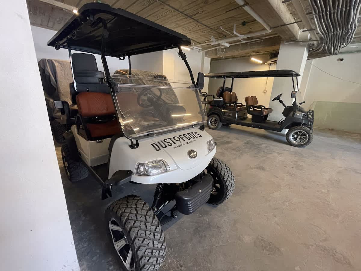 Two golf carts are parked in a concrete garage or covered parking area, with the white cart in the foreground featuring brown quilted seats and large off-road tires.